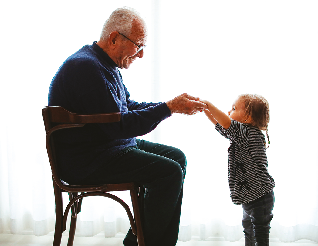 La fotografia riprende un uomo anziano seduto, un nonno, che stringe le mani di una bambina, forse la sua nipotina, aiutandola a stare in piedi. 