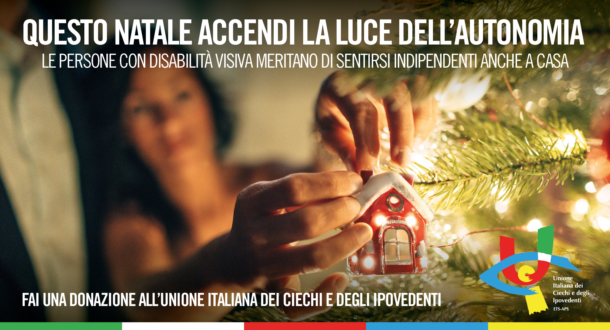 Nell’immagine c’è una figura maschile, appena intuibile nello sfondo sfocato, accanto a una donna illuminata da una luce calda e morbida tipica dell’atmosfera natalizia. In primo piano, le mani di lei appendono una piccola decorazione a forma di casetta rossa su un ramo dell’albero di Natale. La scena trasmette attenzione e la magia discreta di un gesto semplice ma pieno di significato. In alto la scritta: Questo Natale accendi la luce dell'autonomia. Le persone con disabilità visiva meritano di sentirsi indipendenti anche a casa; in basso: Fai una donazione all'Unione Italiana dei Ciechi e degli Ipovedenti accanto al logo UICI.