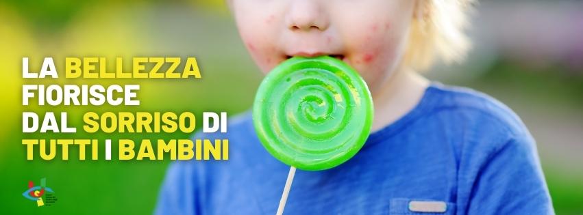 Nellimmagine un Bambino con maglietta blu a maniche corte felice di mangiare un lecca lecca rotondo di colore verde. In giallo  presente la scritta La bellezza fiorisce dal sorriso di tutti i bambini